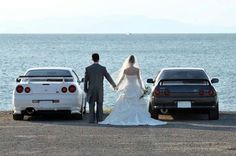 a bride and groom holding hands next to their cars by the water with mountains in the background