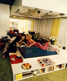 two women sitting on a bed in a room with bookshelves and other furniture