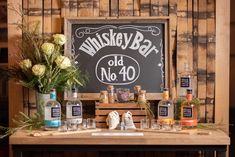 a wooden table topped with lots of bottles and glasses next to a chalkboard sign