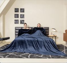 two women sitting on top of a bed covered in a blue blanket and covering them