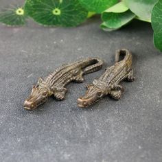 two small alligator figurines sitting next to each other on a stone surface with green leaves in the background