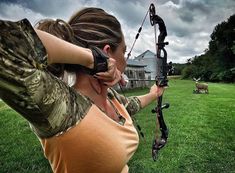 a woman holding a bow and aiming it at an animal in the field behind her