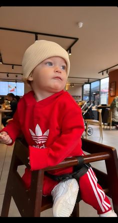 a baby sitting in a wooden chair wearing a red adidas sweater and white hat