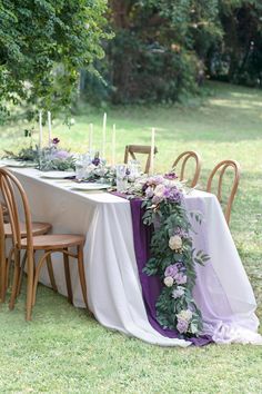 the table is set with purple flowers and greenery on it, along with candles