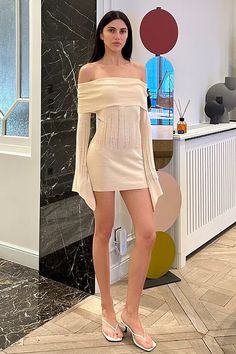 a woman standing in front of a black and white marble wall wearing a beige off the shoulder dress