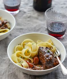 two bowls filled with pasta and meat on top of a table next to wine glasses