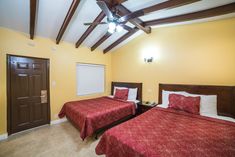 two beds in a room with yellow walls and wood beams on the ceiling, one has red comforters