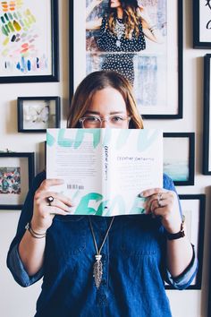 a woman is reading a book in front of pictures on the wall and holding it up to her face