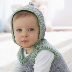 a baby wearing a crochet hooded vest and hat on the floor in front of a bed