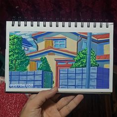 a hand holding up a drawing of a house in front of a fence and trees