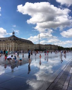 many people are walking around in front of a large building with water spewing from it