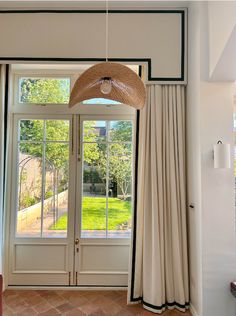 an open door and window in a room with white walls, beige curtains, and a straw hat hanging from the ceiling