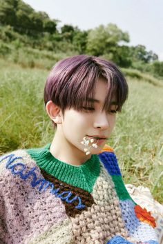 a young man with purple hair is sitting in the grass wearing a sweater and eating something