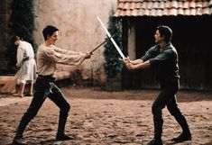 two men are holding swords in the middle of an outdoor area with people standing around