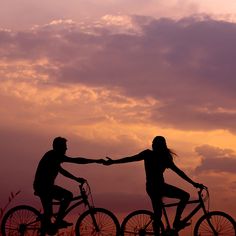 two people on bicycles holding hands in front of an orange and purple sky with clouds