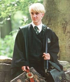 a young boy dressed in a harry potter costume holding a bag and briefcase while standing next to a tree