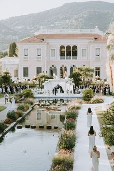 two women in white dresses are walking towards a large pink building with a fountain and palm trees