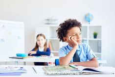 two children sitting at desks with books in front of them and one child holding his hand to his face