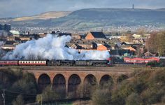 a train traveling over a bridge with steam pouring out of it's stacks and buildings in the background