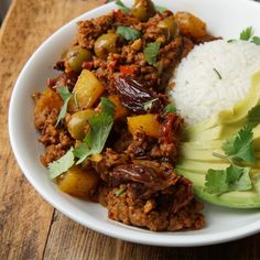 a white plate topped with meat, rice and avocado on top of a wooden table