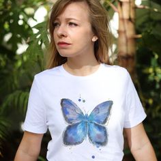 This elegant white t-shirt for women features a stunning butterfly print that has been hand-painted by a talented artist.  The soft and breathable fabric ensures maximum comfort, while the vibrant colors of the print create a bold and eye-catching look.  The design is perfect for adding a touch of elegance and sophistication to any outfit, whether dressed up or down.  Made with high-quality materials and attention to detail, this t-shirt is sure to become a favorite in any wardrobe.  Get yours today and add a unique and artistic touch to your style! Butterfly Fabric Painting, Butterfly Fabric, Womens Graphic Tees, Tshirt Painting, T Shirt For Women, Butterfly Print, Fabric Painting, Graphic Tees Women, White T Shirt