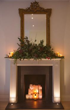 candles are lit in front of a fireplace with an ornate mirror above it and greenery on the mantle