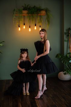 two women in black dresses standing next to each other on a wooden floor with plants hanging from the ceiling