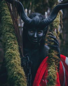 a woman dressed in black with horns and makeup is holding onto moss covered branches while standing in the woods