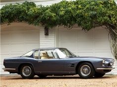 an old blue sports car parked in front of a white house with two garage doors