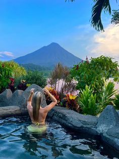 a woman sitting in the water next to some rocks and plants with a volcano in the background