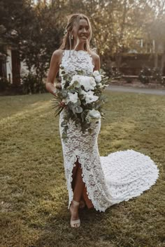 a woman in a white lace dress holding a bridal bouquet standing on the grass