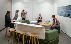 people are sitting at tables in an office with white walls and green chairs, while one woman is writing on the wall