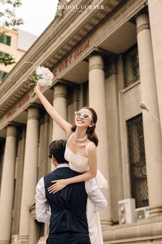 a man and woman standing in front of a building with flowers on their shoulders, one holding the other up