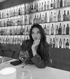 a woman sitting at a table with a glass of wine in front of her and bottles on the wall behind her