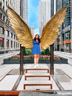 a woman is standing in front of a golden angel statue with her arms raised up