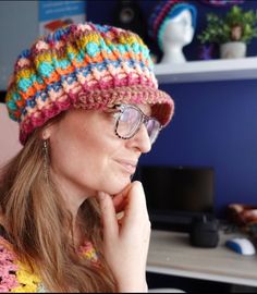 a woman wearing glasses and a multicolored crocheted hat sits in front of a computer desk