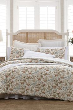 a bed with floral comforter and pillows in a white room next to two windows