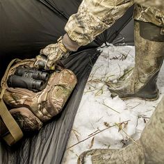 a man standing next to a tent in the snow with his boots and bag on it