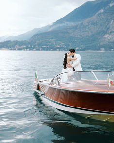 a bride and groom standing on the back of a boat in the water with mountains in the background