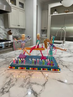 two barbie dolls sitting on top of a birthday cake with candles in the shape of letters