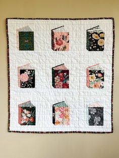 a quilted wall hanging on the side of a wall with flowers and books in it