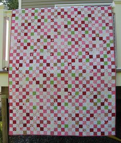 a pink and green checkered quilt hanging on the side of a house with trees in the background