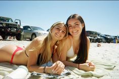 two young women laying on the beach in front of their cars and towels, smiling at the camera