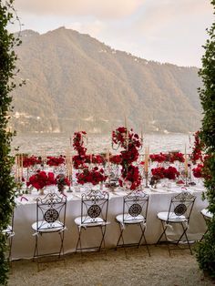 an outdoor table set up with red flowers and greenery for a formal dinner by the water