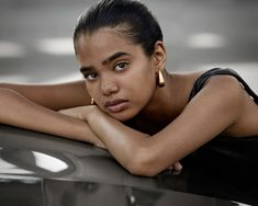 a beautiful young woman leaning on the hood of a car with her arms crossed and looking at the camera