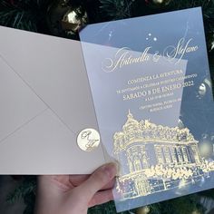 a person holding up a wedding card in front of a christmas tree with lights on it