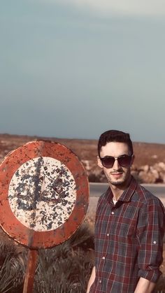 a man wearing sunglasses standing next to a rusted road sign in the middle of nowhere