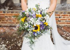 a bride holding a bouquet of sunflowers and greenery in front of a brick wall