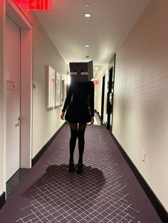 a woman walking down a long hallway with a red exit sign on the wall behind her