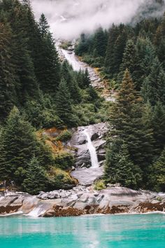 a waterfall in the middle of a forest with blue water and trees on both sides